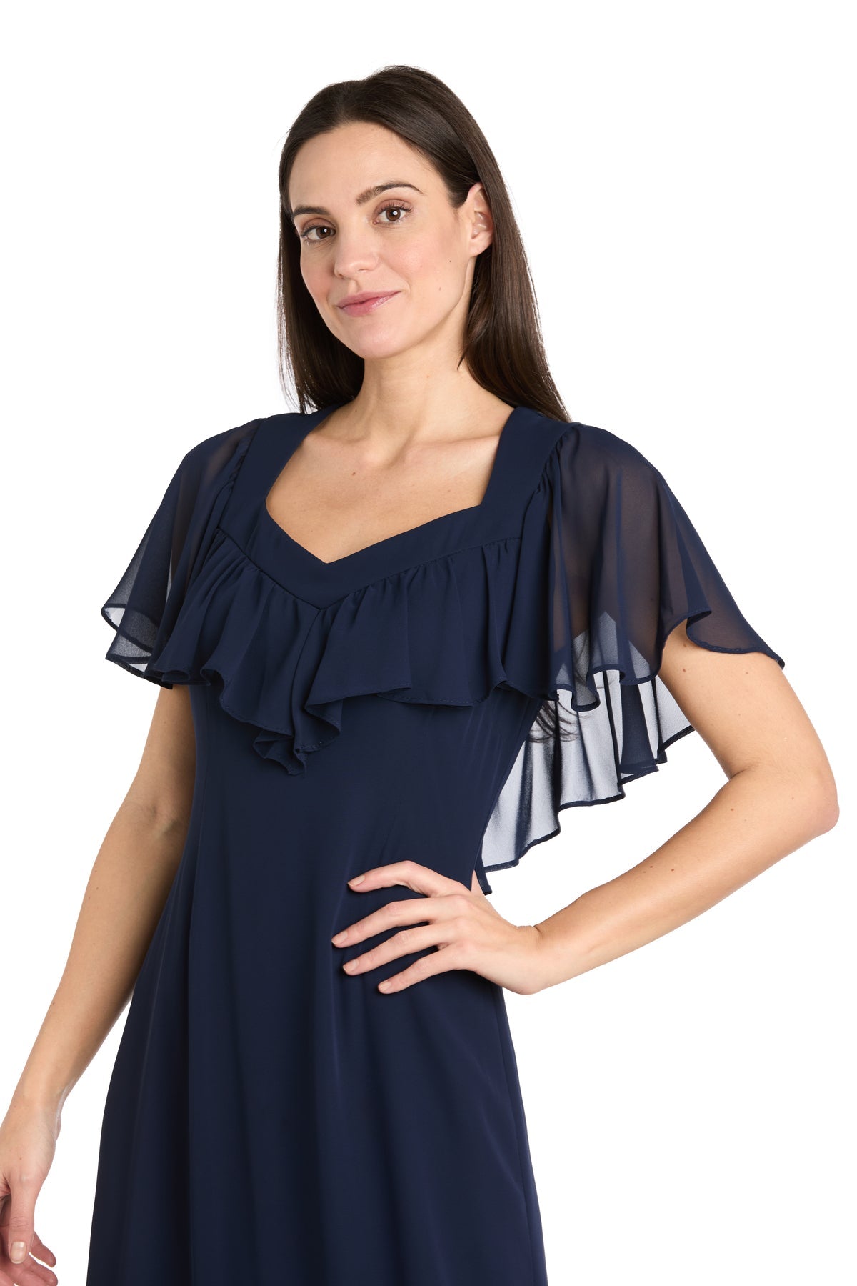 Woman modeling the R&M Richards 4054P Petite Navy Blue Midi Party Dress with sheer, ruffled sleeves, posing with one hand on her hip against a white background.